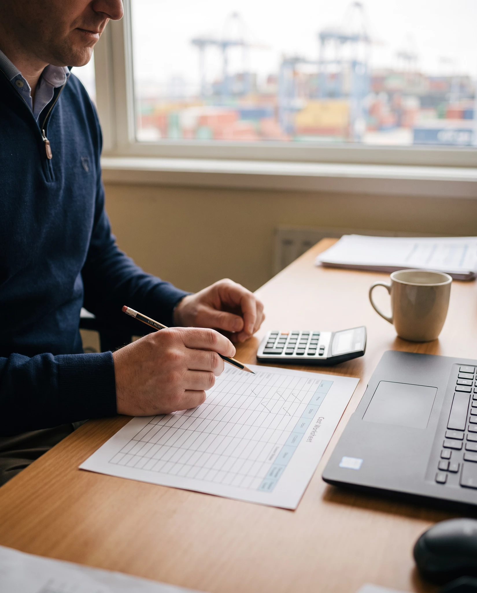 Man in navy quarter-zip sweater filling in a grid worksheet with a pencil at a wooden desk, calculator, laptop