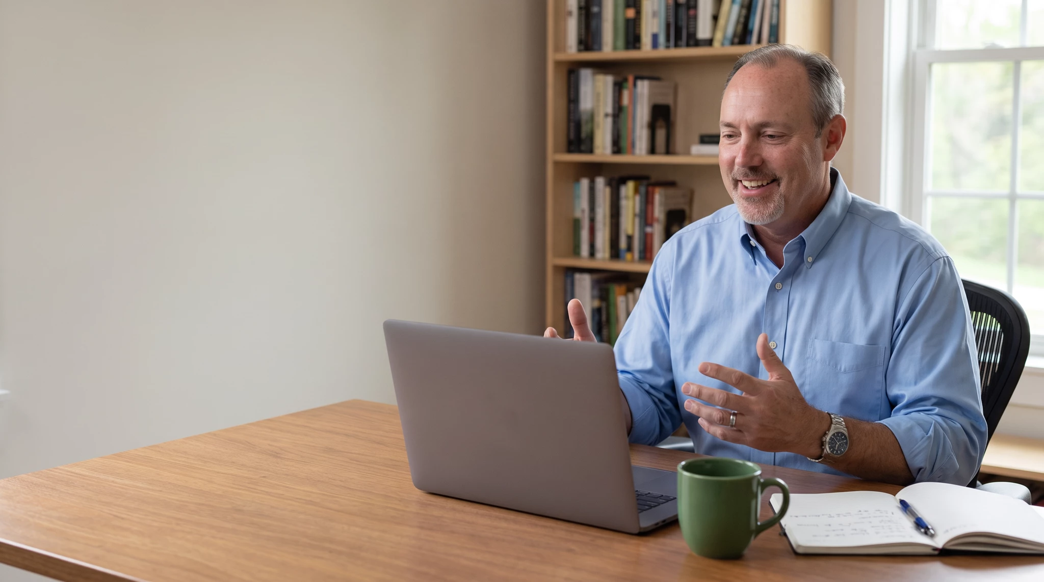 Smiling man in blue button-down gesturing while talking on a laptop video call at a wooden desk, bookshelf and window