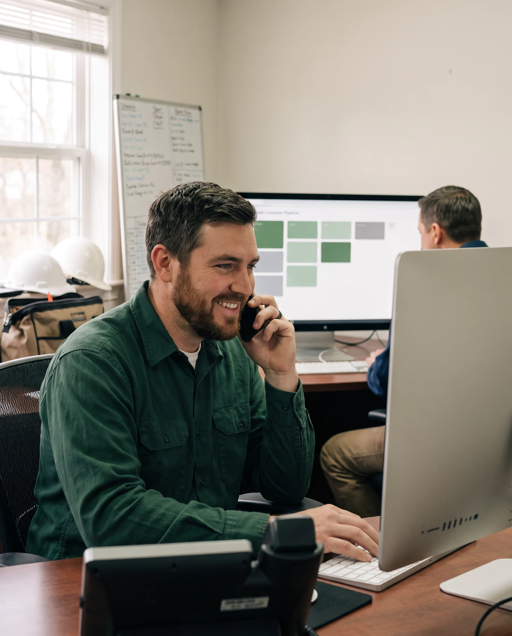 Smiling man in green work shirt talking on a phone at a desk with an iMac