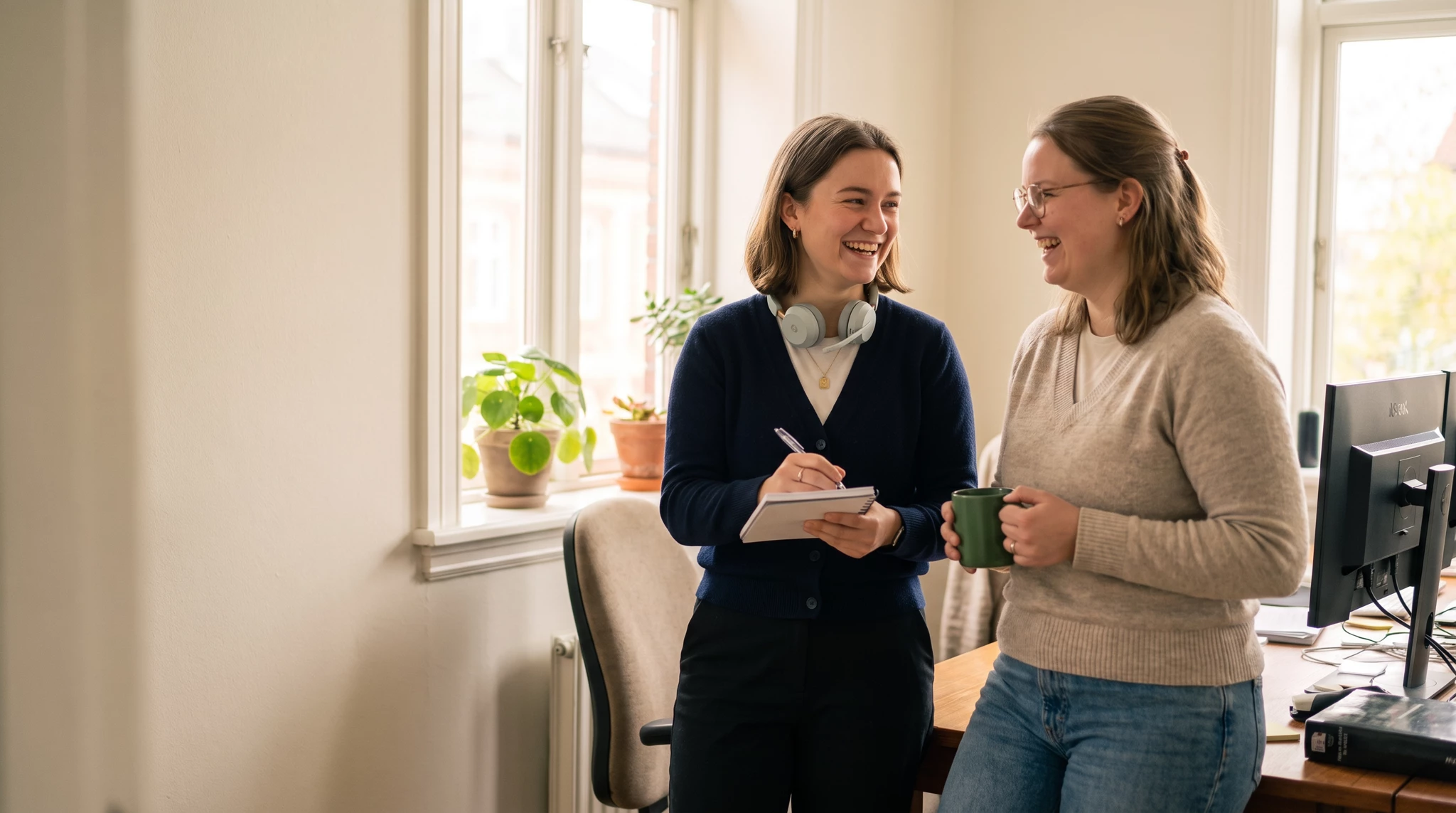Two smiling women chatting in a bright office, one holding a notepad and pen with headphones around her neck